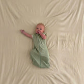 Baby in a sage green sleep sack lying on a beige fabric cover