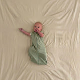 Baby in a sage green sleep sack lying on a beige fabric cover