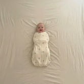 Baby in a warm white 1.0 TOG swaddle on a beige blanket